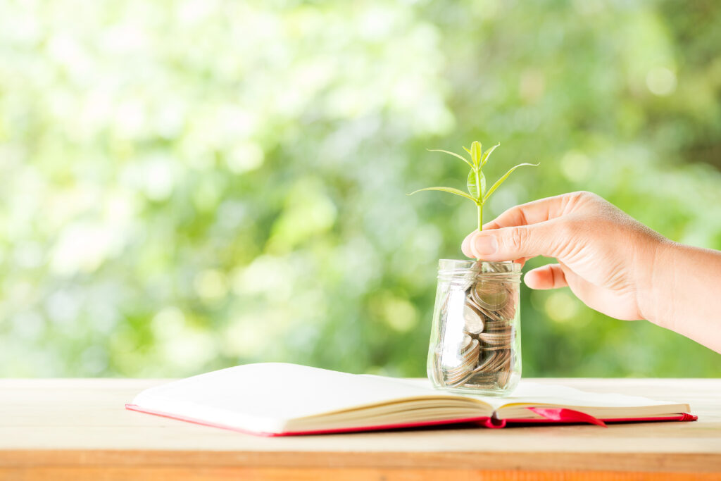 A hand placing a small green plant into a glass jar filled with coins on an open notebook representing easy passive income ideas for beginners